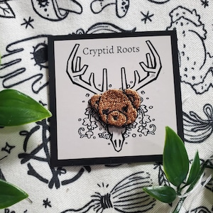 May include: A brown embroidered bear head patch with a skull and antlers, on a white card with the text "Cryptid Roots".