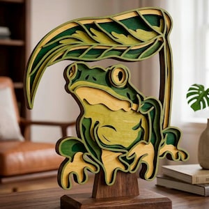 May include: A layered wooden frog sculpture with a leaf overhead. The frog is primarily green and yellow, with a dark brown base. The leaf and frog are made of multiple layers of wood, creating a 3D effect. The sculpture is a decorative item.