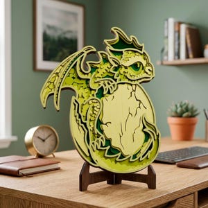 May include: A layered wooden dragon sculpture in shades of green and yellow, emerging from an egg. The dragon is mounted on a dark brown wooden stand, with intricate details visible. The sculpture is displayed on a wooden desk.