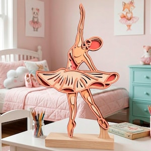 May include: A layered wooden ballerina sculpture in shades of peach and orange, posed mid-dance. The sculpture is mounted on a rectangular wooden base. The ballerina is in a tutu, with arms raised. The background includes a child's bedroom with pink decor.