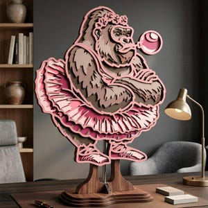 Könnte beinhalten: Mehrschichtige Holzskulptur eines Gorillas mit rosa Tutu und Blumenkranz, der eine Blase bläst. Die Skulptur in Braun- und Rosatönen ist auf einem Holzsockel montiert. Ein skurriles und künstlerisches Wohnaccessoire.