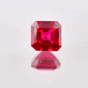 May include: A deep red gemstone with a square cut and a polished finish. The gemstone is reflecting light, creating a bright red glow.