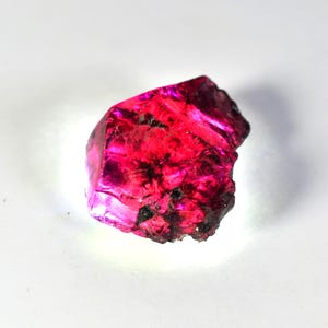 May include: A raw, uncut gemstone with a vibrant, deep pink hue. The crystalline structure is visible, with sharp edges and a rough, textured surface. Small dark inclusions are scattered throughout the stone, adding to its natural beauty.
