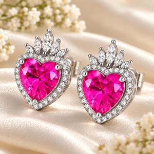 May include: A pair of silver-toned stud earrings featuring a large, heart-shaped, vibrant pink center stone. Each earring is surrounded by a halo of small, clear, round-cut stones and topped with a crown-like arrangement of marquise-cut stones.