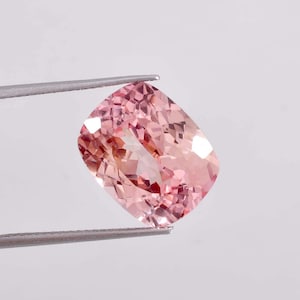 Op de afbeelding: Een roze edelsteen met een kussen-slijpsel, vastgehouden door een pincet tegen een witte achtergrond.