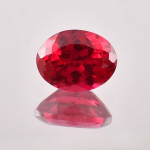 May include: A deep red oval-shaped gemstone with a faceted cut. The gemstone is reflecting light and has a shiny finish.