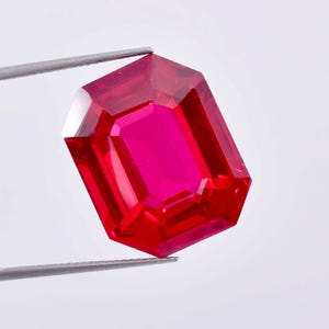 Op de afbeelding: Een levendige rode smaragd geslepen edelsteen vastgehouden door een pincet. De edelsteen is een diepe, rijke rode kleur en heeft een hoge mate van helderheid.