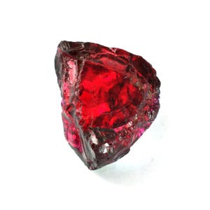 May include: A raw, uncut gemstone with a deep red hue. The gem has a rough, irregular shape and a translucent quality, allowing light to pass through. The surface has a slightly glossy appearance, reflecting light. The gemstone is set against a white background.