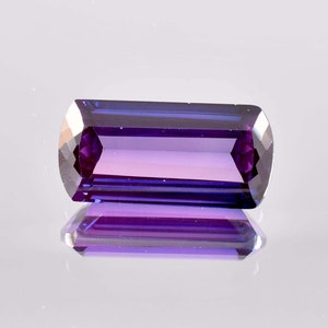 Può includere: Una gemma di colore viola intenso con taglio rettangolare e finitura sfaccettata. La gemma riflette la luce, mostrando la sua brillantezza.