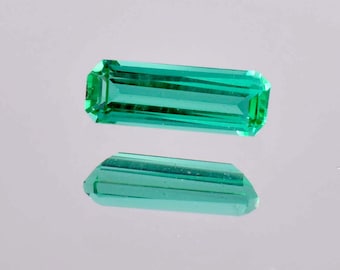 May include: A vibrant green gemstone with a rectangular shape and a faceted cut. The gemstone is displayed on a reflective surface.