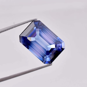 以下が含まれることがあります： 長方形のエメラルドカットの濃い青色の宝石。宝石は白い背景に対してピンセットで保持されています。