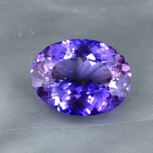 Puede incluir: Una gran gema de amatista de forma ovalada, de color morado oscuro. La gema tiene un corte facetado y está colocada sobre un fondo gris.