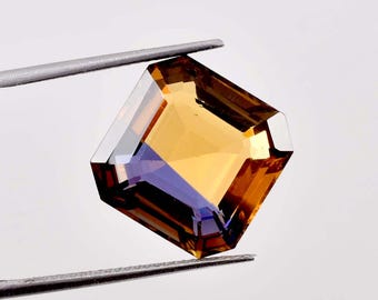 May include: A faceted gemstone held by tweezers. The gem has a square shape with beveled edges. The colors are a mix of amber, gold, and purple, with a high degree of clarity. The gem is likely a cut stone for jewelry.