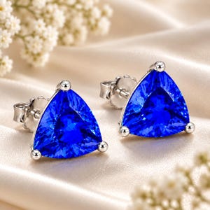 May include: A pair of stud earrings featuring vibrant, triangular-cut blue gemstones set in polished silver-toned metal. Each earring has a three-prong setting that secures the faceted stone, complemented by a standard post and butterfly back closure.