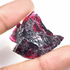 May include: A rough, uncut gemstone with a deep red hue, held between two fingers. The stone has a jagged, irregular shape and a semi-transparent quality, with areas of dark and light red. The overall appearance suggests a natural, unpolished mineral specimen.