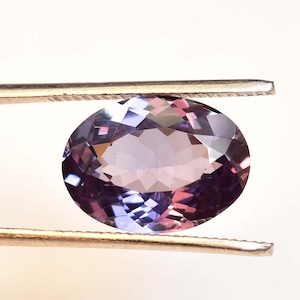 May include: A purple oval-shaped gemstone with facets, held in tweezers against a white background.