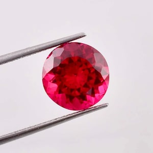 May include: A round, faceted, red gemstone held by tweezers. The gemstone is a deep, rich red color and has a high level of brilliance.