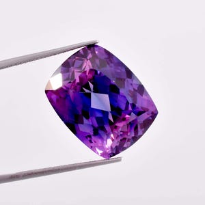 以下が含まれることがあります： 深い紫色の色合いで、縁が薄いピンク色の大きなクッションカットのアメジスト。宝石はピンセットで固定されています。