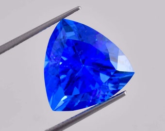 Può includere: Una gemma blu vibrante con taglio triangolare, tenuta da una pinza su uno sfondo bianco.
