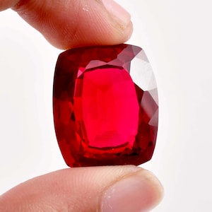 May include: A deep red gemstone with a cushion cut. The gemstone is held in a hand.