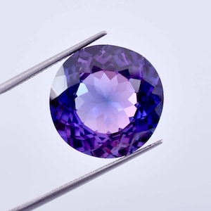 Può includere: Una pietra preziosa di ametista rotonda e sfaccettata con un gradiente da viola a rosa. La pietra preziosa è tenuta da una pinzetta su uno sfondo bianco.