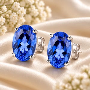 May include: A pair of stud earrings featuring large, oval-cut, vibrant blue gemstones set in polished silver-toned metal. Each earring is secured with a four-prong setting and includes a standard post and butterfly clutch backing mechanism.