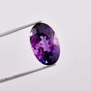 Può includere: Una pietra preziosa ovale di colore viola con taglio a faccette. La pietra preziosa è tenuta da una pinzetta su uno sfondo bianco.