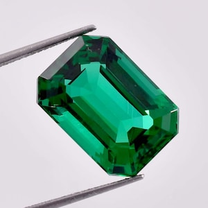 May include: A vibrant green emerald gemstone, cut in an emerald shape, is held by tweezers. The gemstone is clear and has a brilliant shine.