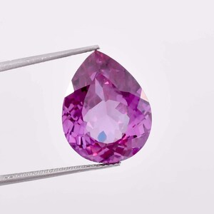 Può includere: Una pietra preziosa a forma di pera con una tonalità viola intenso. La pietra preziosa è tenuta da una pinzetta su uno sfondo bianco.