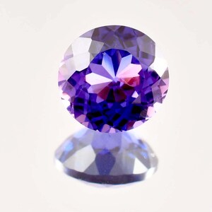 May include: A faceted, oval-cut gemstone with a deep purple hue. The gem reflects light, showcasing a complex pattern of internal facets. The stone is set against a white background, with its reflection visible below.