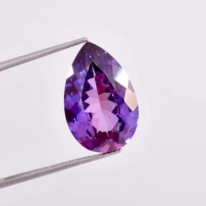 Op de afbeelding: Een peervormige amethist edelsteen met een paarse tint en facetten, vastgehouden door een pincet.
