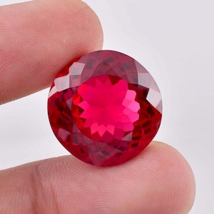 May include: A large, round, red gemstone with a faceted cut. The gemstone is held in a hand.