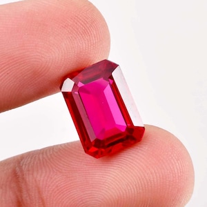 Può includere: Una gemma di colore rosso vibrante, taglio smeraldo, che mostra una tonalità profonda e ricca. La gemma è tenuta in una mano, evidenziando le sue dimensioni e la sua brillantezza.