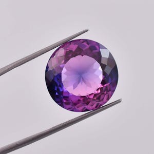 Può includere: Una gemma rotonda e sfaccettata con un gradiente da viola a rosa. La gemma è tenuta da una pinza su uno sfondo bianco.