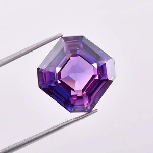 Può includere: Una gemma sfaccettata, a taglio quadrato, con un centro viola intenso e sfumature sfumate di viola e blu. La gemma è tenuta da pinzette d'argento, che ne evidenziano il design simmetrico e le brillanti sfaccettature. La gemma è probabilmente per la creazione di gioielli.