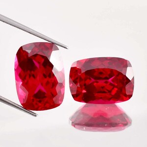 May include: Two red cushion-cut gemstones with a faceted finish. The gemstones are displayed on a white surface.