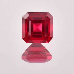 Puede incluir: Una gema roja vibrante con un corte cuadrado esmeralda. La gema está facetada y refleja la luz.