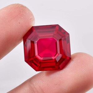 May include: A deep red gemstone with a square shape and a faceted cut. The gemstone is held in a hand.