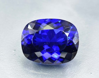 Può includere: Una gemma sfaccettata, taglio a cuscino, di colore blu intenso. Le sfaccettature della gemma riflettono la luce, creando un effetto brillante. La forma è un rettangolo arrotondato, ideale per gioielli. La gemma è su uno sfondo chiaro.