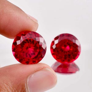 May include: Two round, red gemstones with a faceted cut. The gemstones are on a white surface.
