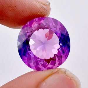 May include: A round, faceted amethyst gemstone with a purple and pink hue. The gemstone is held in a hand.