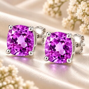 May include: A pair of stud earrings featuring cushion-cut, vibrant purple gemstones set in polished silver-toned metal. Each earring is secured with a matching metal post and butterfly back, showcasing a faceted surface that reflects light.