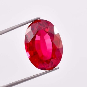 Puede incluir: Una gema ovalada de color rojo vibrante con un corte facetado. La gema está sujeta por unas pinzas.