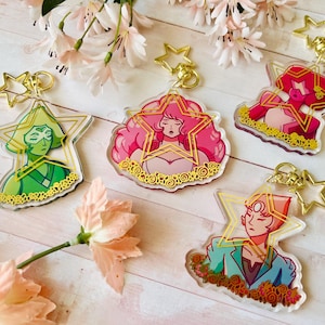 May include: Five character keychains with gold-colored star-shaped hardware. Each keychain features a different character design with vibrant colors: green, pink, and red. The keychains are on a light wood surface with pink flowers.