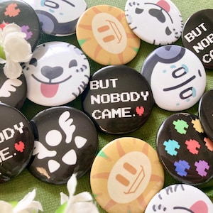 Puede incluir: Una colección de botones redondos y coloridos con varios diseños. Algunos botones presentan el texto "BUT NOBODY CAME" en píxeles. Otros botones tienen caras de dibujos animados, calaveras y patrones abstractos. Los botones están esparcidos sobre una superficie verde.