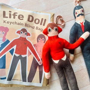 May include: A brown paper Life Doll Keychain Blind Bag with a printed illustration of three characters, accompanied by two fabric doll keychains. One doll wears a red sweater and brown trousers, while the other wears a dark suit with a red tie.