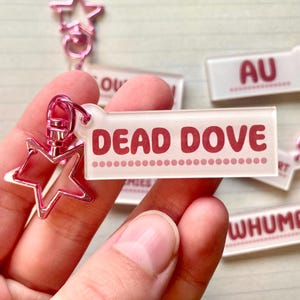 May include: A clear acrylic keychain with the words "DEAD DOVE" in red letters. The keychain has a pink star-shaped clasp and a pink star charm. Other keychains with text are visible.