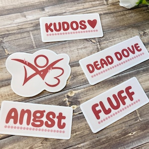 May include: Five white stickers with red text and designs. The stickers include the words "KUDOS" with a heart, "DEAD DOVE", "FLUFF", "Angst", and a stylized figure. The stickers are on a wooden surface.