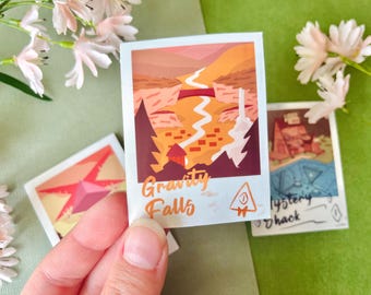 Könnte beinhalten: Eine Hand hält drei quadratische Aufkleber mit Illustrationen. Der Aufkleber in der Mitte zeigt "Gravity Falls" mit einer Landschaftsszene. Die anderen Aufkleber zeigen einen Stern und eine Figur aus der Serie. Die Aufkleber befinden sich auf einer grünen Oberfläche mit Blumen.