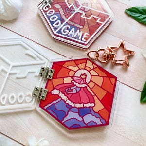 Puede incluir: Llaveros acrílicos con un diseño de vidriera que presenta una figura con capa y corona, con las palabras "GOOD GAME". También se ve un cierre en forma de estrella de oro rosa.